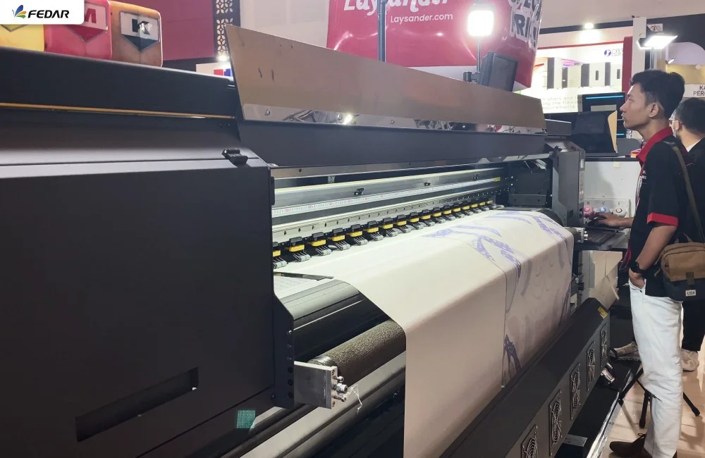 Mesin printer industri berukuran besar sedang beroperasi di area pameran Surabaya Printing Expo 2026 dengan pengunjung yang menyaksikan demonstrasi langsung.